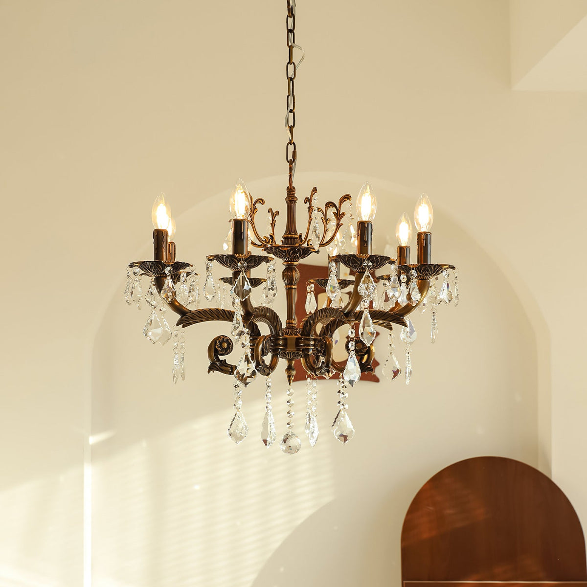 A Clérachic chandelier with bronze scrolled arms and clear prisms hangs from a chain before an arched cream wall, bulbs lit.