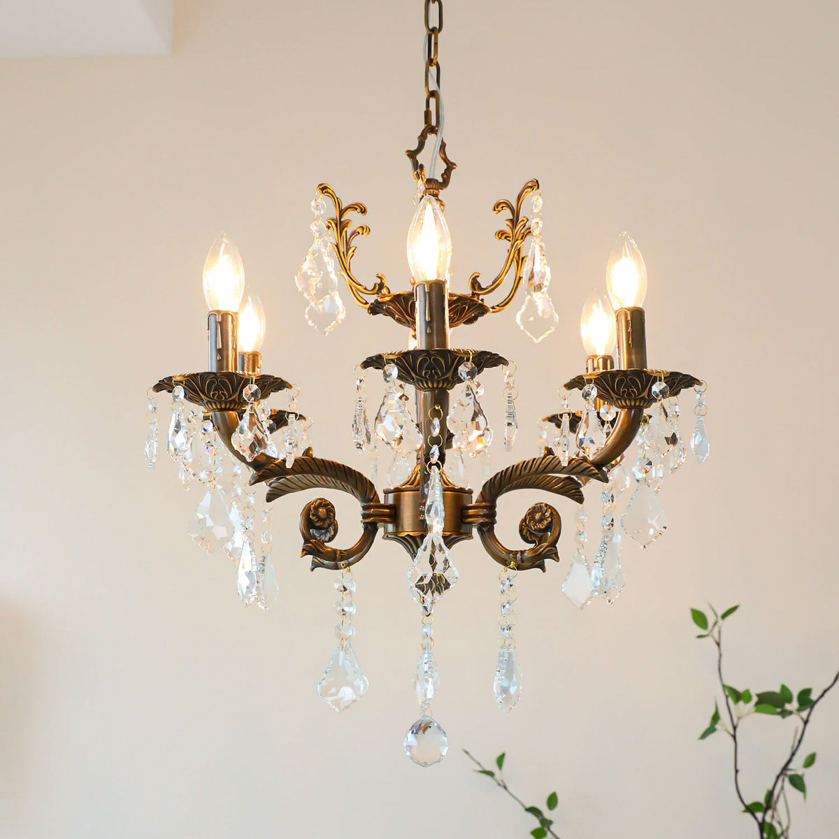 A lit bronze Clérachic chandelier with five candle bulbs and hanging clear crystal drops, with green twigs at the right.