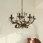 A bronze Clérachic chandelier with clear prisms hangs in front of an arched white wall, with a brown ceramic piece below.
