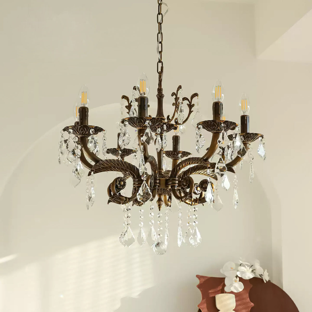 A bronze Clérachic chandelier with clear prisms hangs in front of an arched white wall, with a brown ceramic piece below.