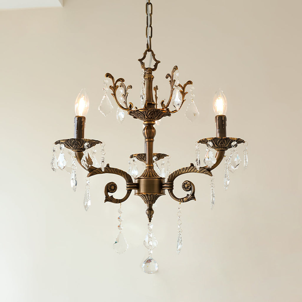 A three-light bronze Clérachic chandelier with scrolled arms and clear prism drops hangs against a plain white wall.