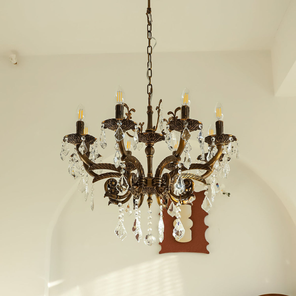 A bronze Clérachic chandelier with eight arms and clear prisms hangs before an arched wall, with a brown mirror behind.