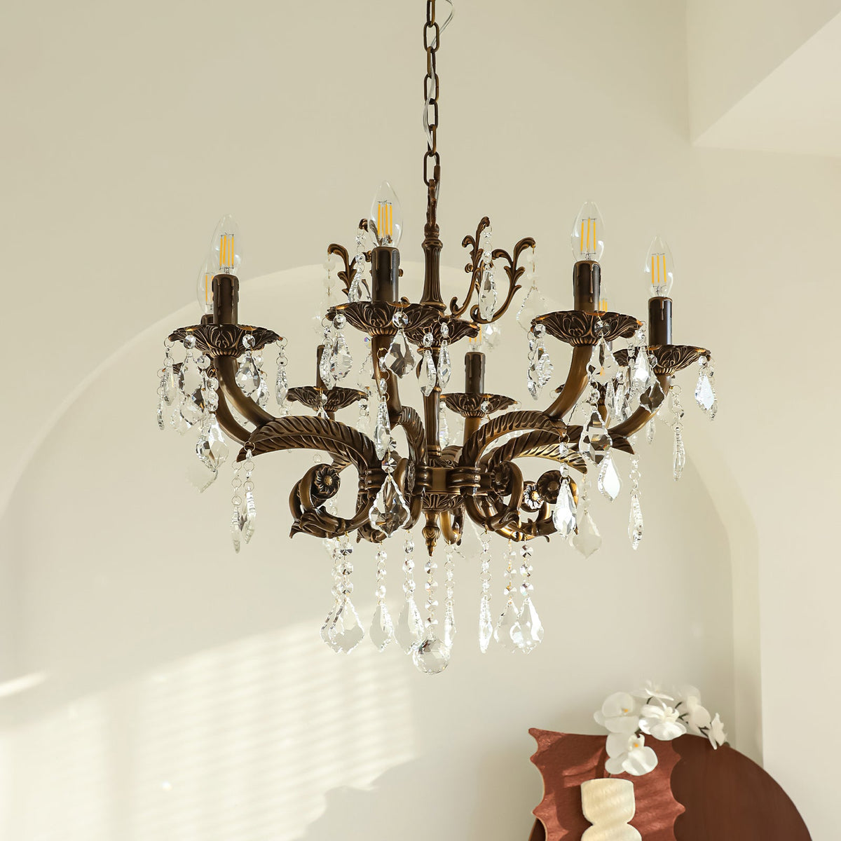 A Clérachic chandelier with bronze scrolled arms and many clear prisms hangs in a pale room beside an arched wall.