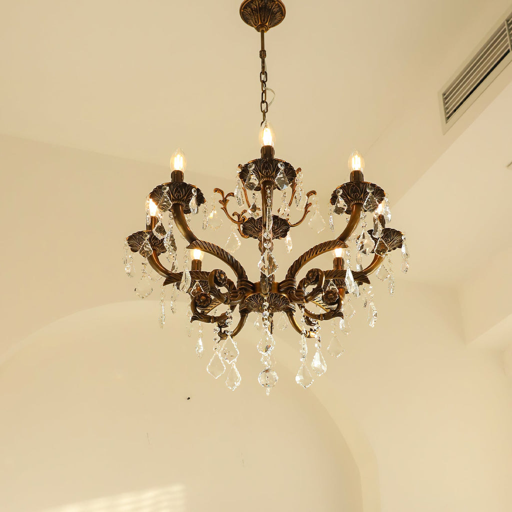 A bronze Clérachic chandelier seen against a warm cream arch, its candle bulbs lit and clear prisms hanging below.