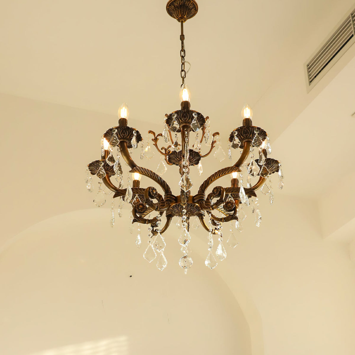 A bronze Clérachic chandelier seen against a warm cream arch, its candle bulbs lit and clear prisms hanging below.