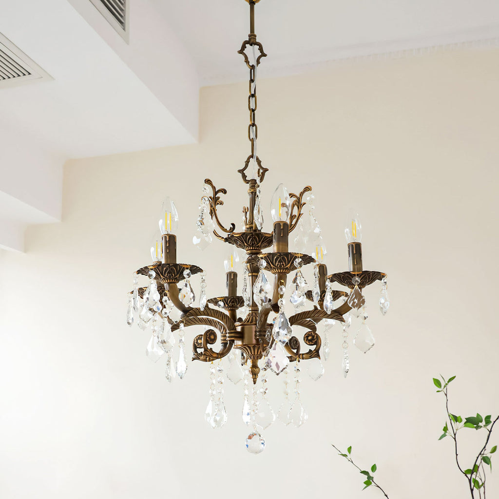 A Clérachic chandelier with bronze arms and clear prisms hangs near a white ceiling recess, with green twigs at the right.