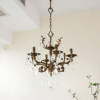 A Clérachic chandelier with bronze arms and clear prisms hangs near a white ceiling recess, with green twigs at the right.