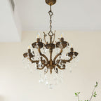 A bronze Clérachic chandelier with a decorative crown, scrolled arms and clear prisms, with a leafy branch behind.