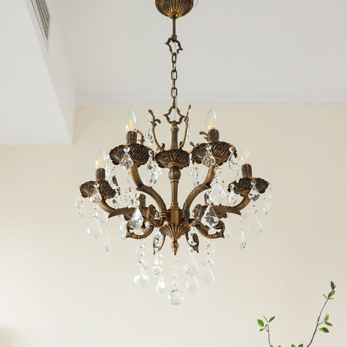 A bronze Clérachic chandelier with a decorative crown, scrolled arms and clear prisms, with a leafy branch behind.
