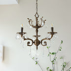 A three-arm bronze Clérachic chandelier with candle bulbs and clear prisms hangs in front of a soft white wall.