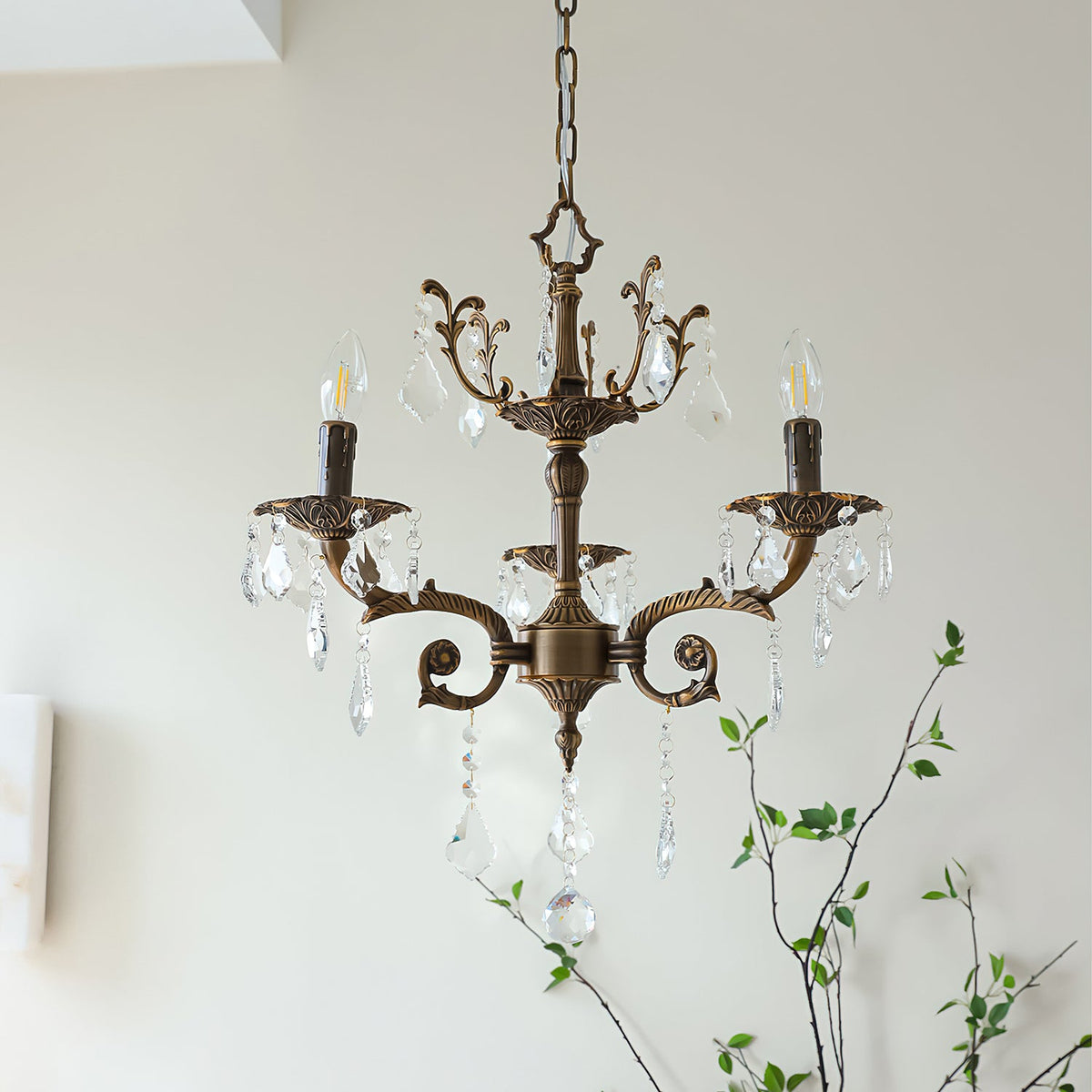 A three-arm bronze Clérachic chandelier with candle bulbs and clear prisms hangs in front of a soft white wall.