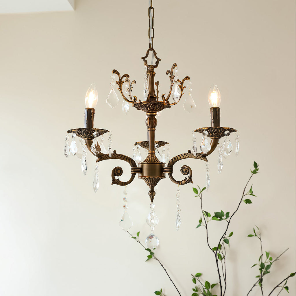 A Clérachic chandelier with three bronze arms and clear prisms, lit, with a leafy branch rising behind it.
