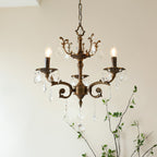 A Clérachic chandelier with three bronze arms and clear prisms, lit, with a leafy branch rising behind it.
