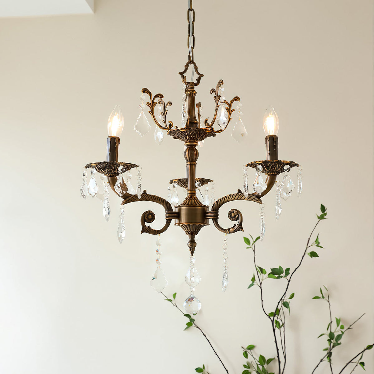 A Clérachic chandelier with three bronze arms and clear prisms, lit, with a leafy branch rising behind it.