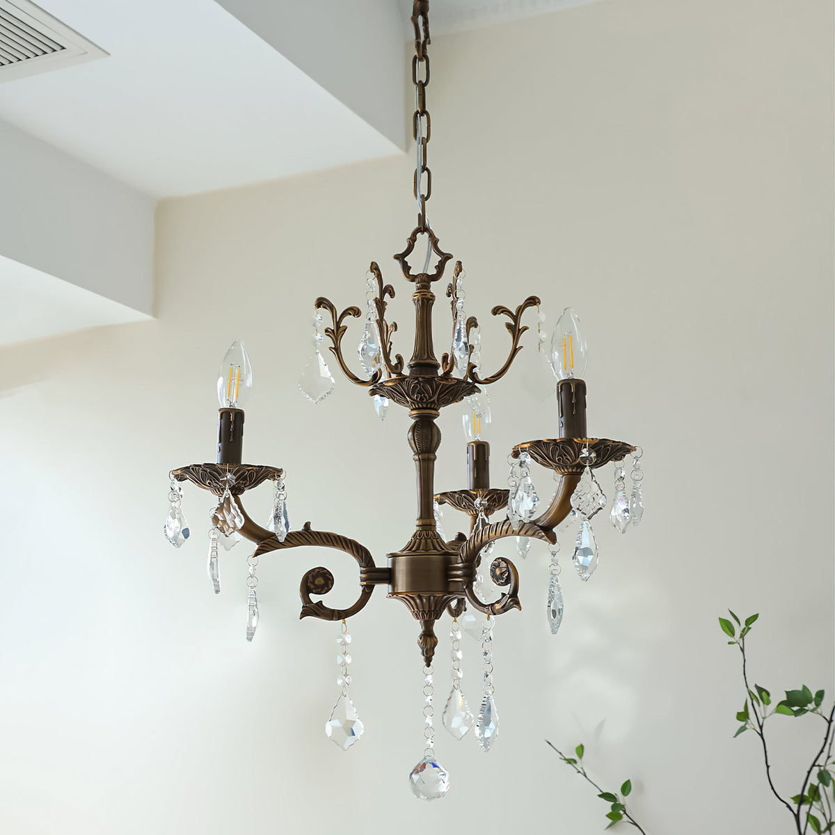 A bronze Clérachic chandelier with three scrolled arms hangs below a white ceiling step, with green twigs behind.