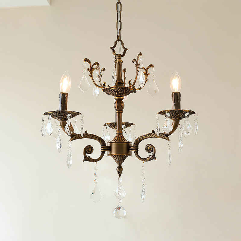 A three-light bronze Clérachic chandelier with an ornate crown and clear prisms hangs against a pale wall, lit.