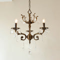 A three-light bronze Clérachic chandelier with an ornate crown and clear prisms hangs against a pale wall, lit.