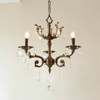 A three-light bronze Clérachic chandelier with an ornate crown and clear prisms hangs against a pale wall, lit.
