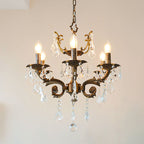 A bronze Clérachic chandelier with six lit candle bulbs and many clear prisms hangs against a soft pink wall.
