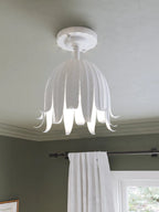 Close view of the Clérachic white petal ceiling light, its slim curled tips lit from inside, above a green wall and curtain.