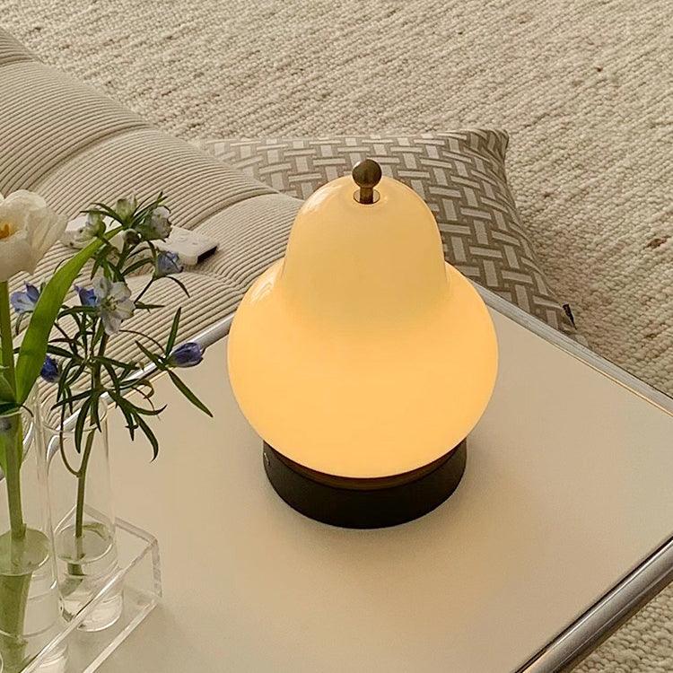 Lit Clérachic pear lamp glowing warm on a pale tray table, beside a glass vase of blue flowers and a cushion on a woven throw.