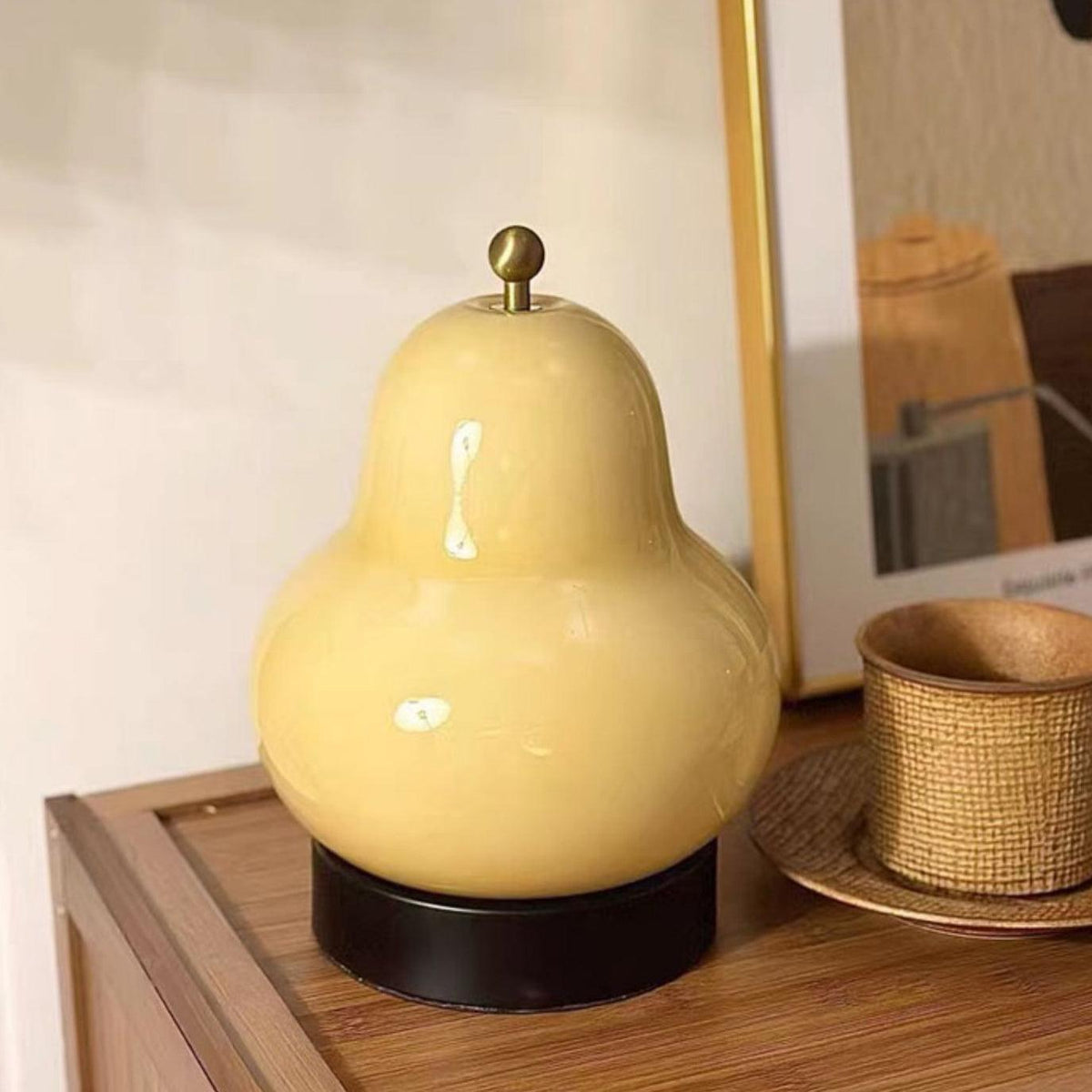This Clérachic pear-shaped lamp in cream glass sits on a wooden bedside shelf, with a framed print and a woven cup on a saucer beside it.