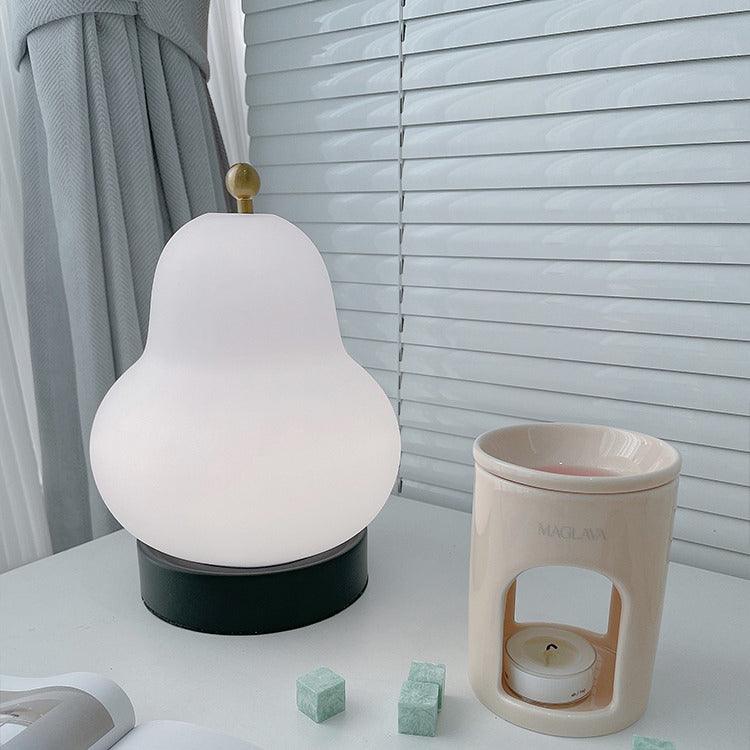 White glass Clérachic pear lamp on a desk by a window with blinds, next to a candle warmer and two small green cubes.