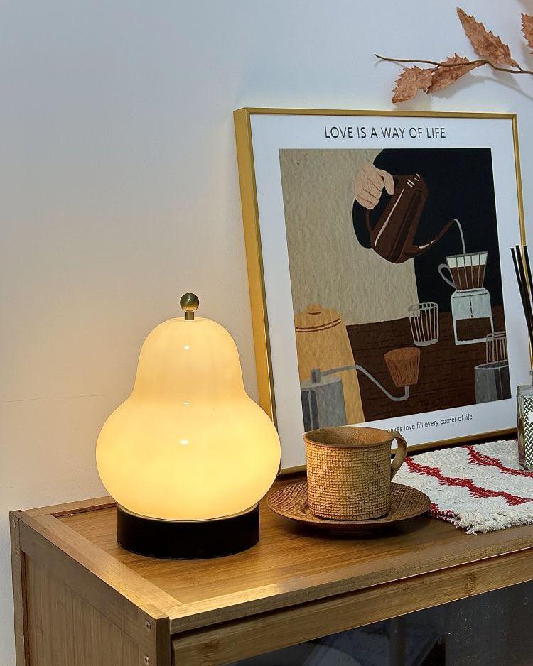 Lit cream Clérachic pear lamp on a bamboo side table, in front of a framed coffee illustration, with a cup on a saucer at its side.