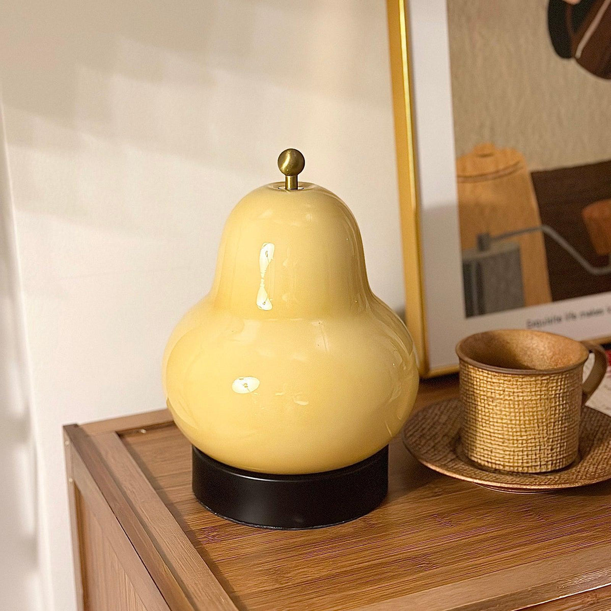Cream Clérachic pear lamp on a wooden cabinet beside a woven cup on a saucer, in front of a gold-framed print.