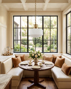 A Clérachic pendant with a brass chain hangs in front of a black-framed window above a round table in a cushioned breakfast nook.