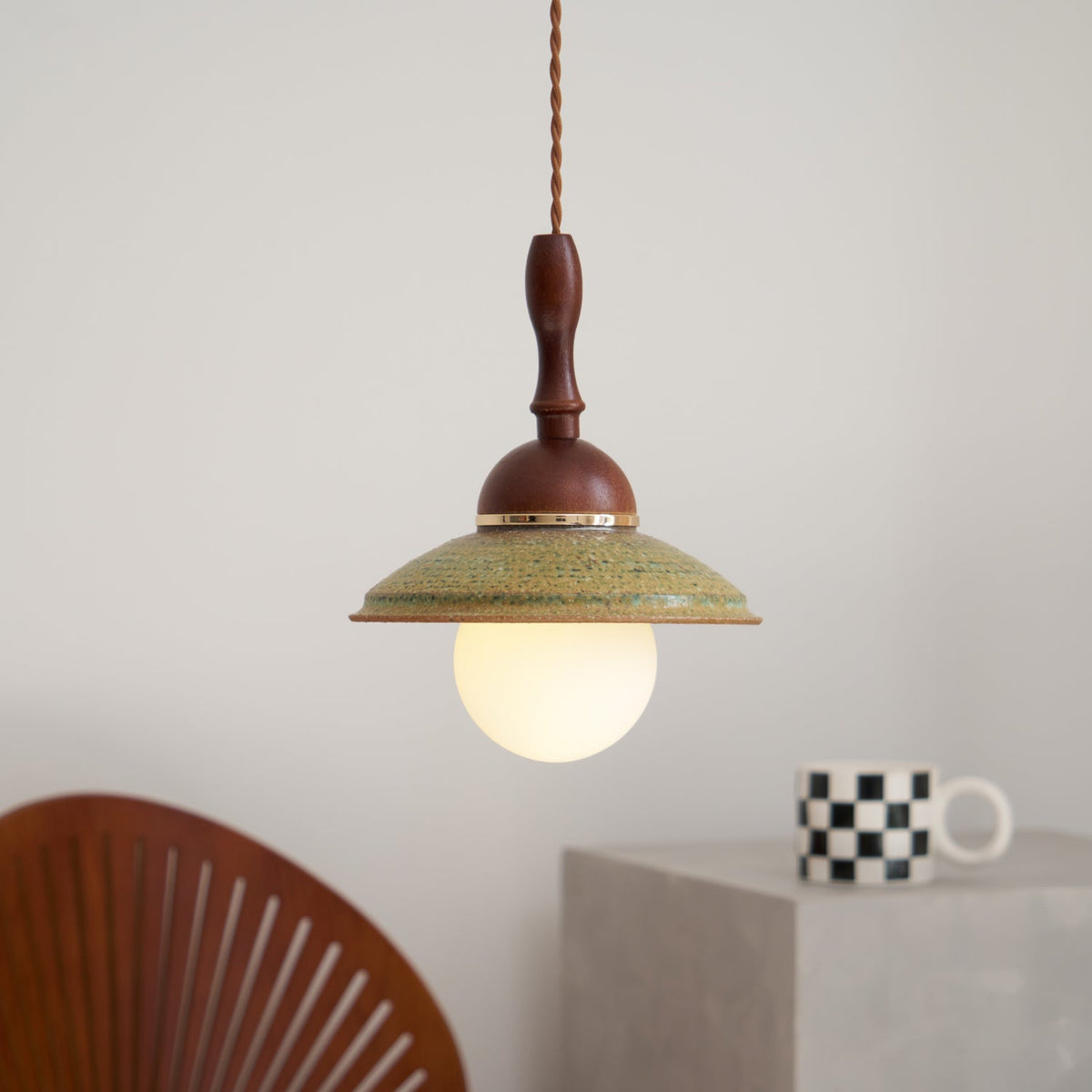 Lit opal globe glowing under the green ceramic shade of this Clérachic pendant, beside a checkered mug and a slatted wooden chair.