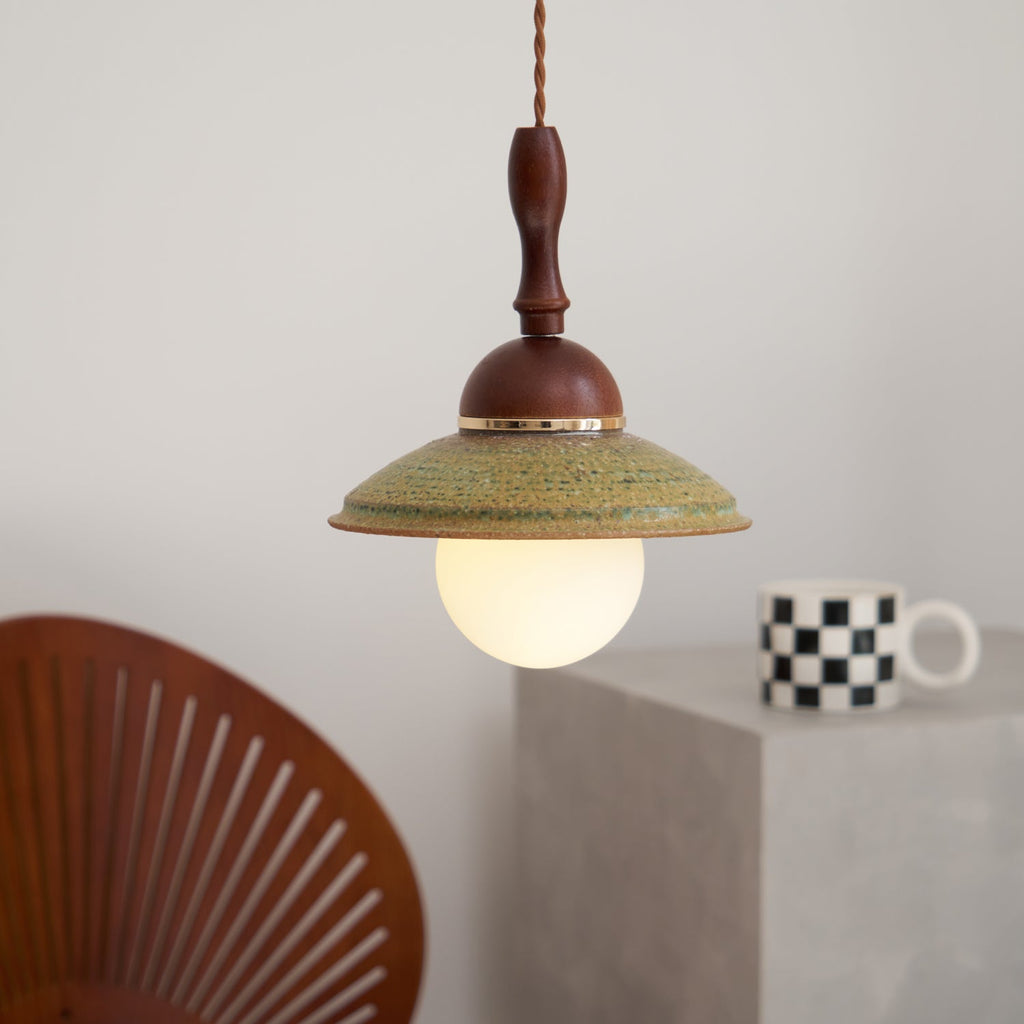 Clérachic pendant with a glowing opal globe, in front of a grey wall with a chequered mug and a rust-red slatted chair.