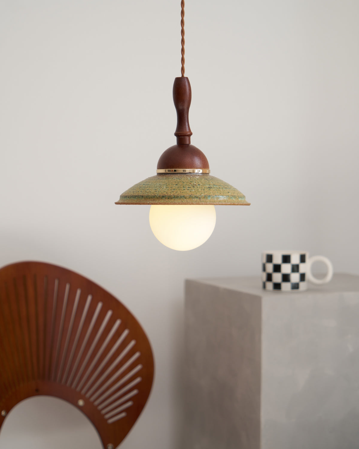 Smaller view of a lit Clérachic pendant, its green shade and globe hanging above a grey block and a slatted chair back.