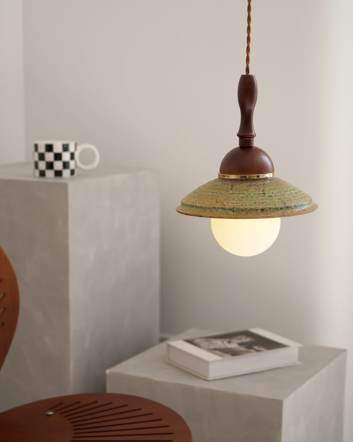 Clérachic pendant hung to one side of the frame, its green shade over a lit globe, with a mug and a book on grey blocks.