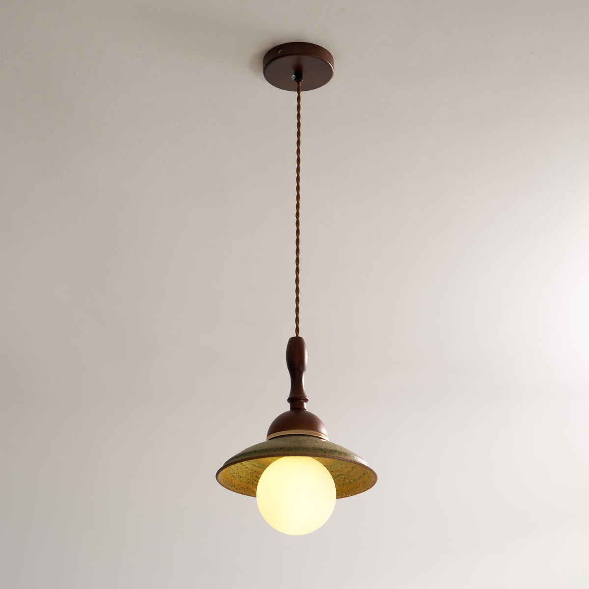Clérachic pendant on its long twisted cord from a round walnut-coloured ceiling canopy, with the globe lit below the green shade.