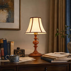 Lit Clérachic table lamp in walnut on a dark wood desk, among stacked books, a blue mug and a small lacquered box, with heavy curtains behind.