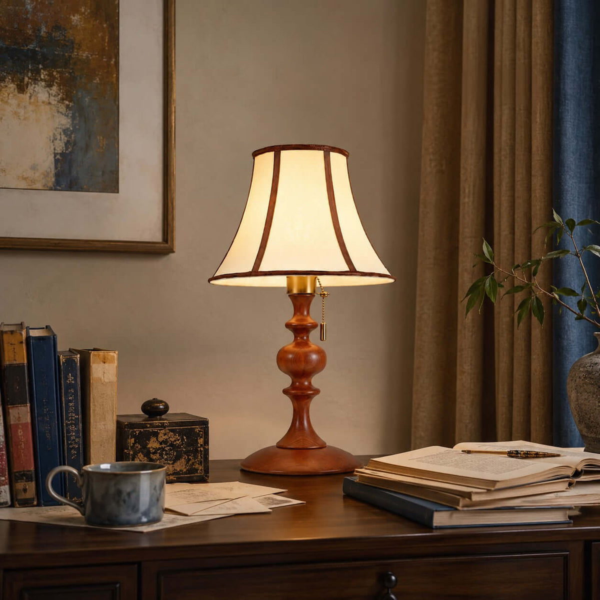 Lit Clérachic table lamp in walnut on a dark wood desk, among stacked books, a blue mug and a small lacquered box, with heavy curtains behind.