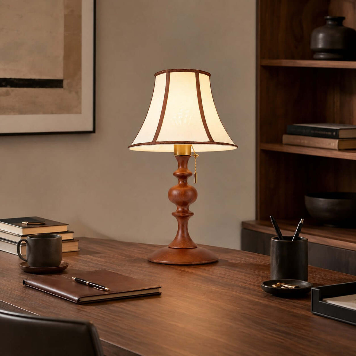 The walnut Clérachic lamp lit on a wooden desk, with a leather notebook, a mug and a pen pot, below a framed print.