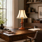Walnut Clérachic table lamp on a leather-topped desk by a window, with bookshelves behind and a vase of green stems.