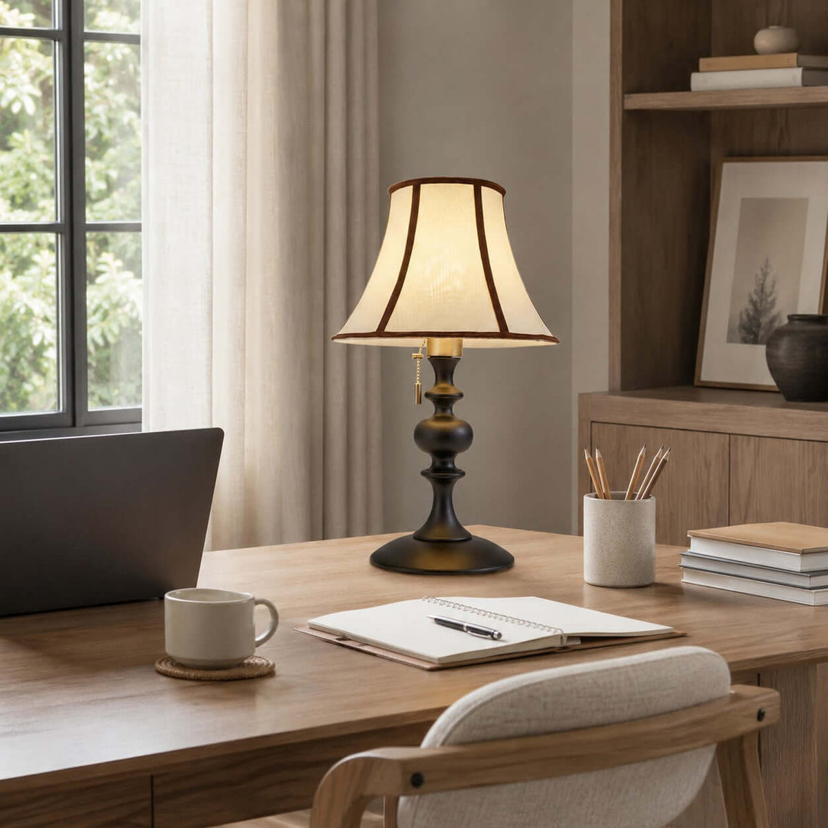 Black-base Clérachic lamp on a light wooden desk beside an open notebook, a mug and a laptop, with a pale chair in front.