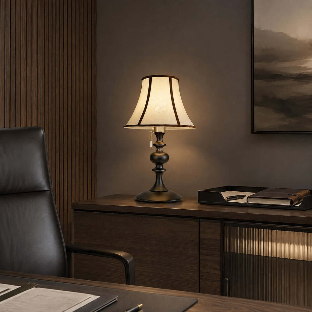 A Clérachic lamp with a black base on a dark wooden sideboard, with a leather desk chair and a dark landscape print behind.