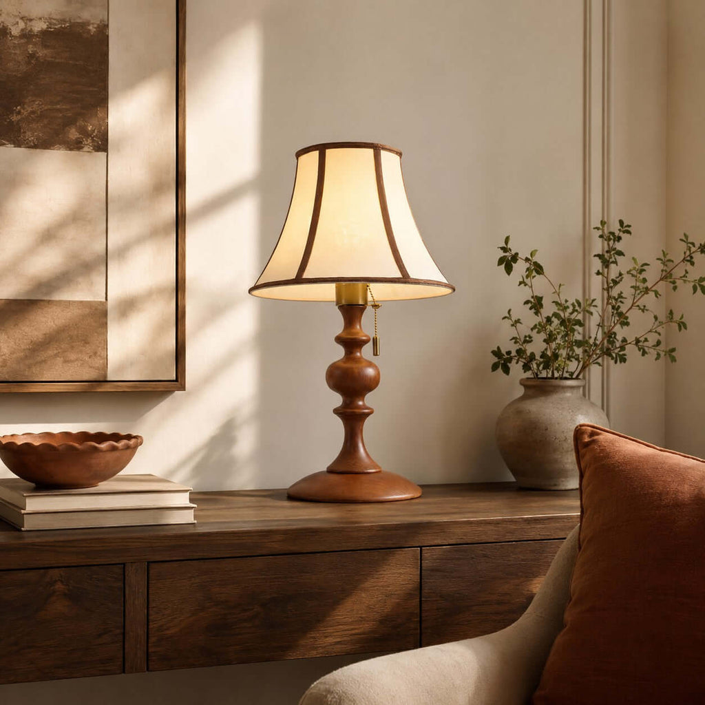 Walnut Clérachic lamp lit on a dark wood sideboard, with a wavy wooden bowl, a stone vase of green stems and a rust cushion.