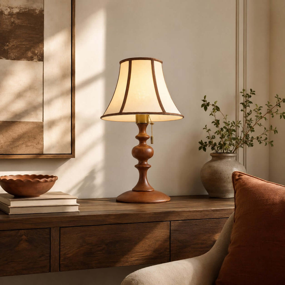 Walnut Clérachic lamp lit on a dark wood sideboard, with a wavy wooden bowl, a stone vase of green stems and a rust cushion.
