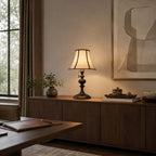 Black-base Clérachic lamp lit on a dark sideboard under a large abstract print, with a small ceramic vase and a window to the left.
