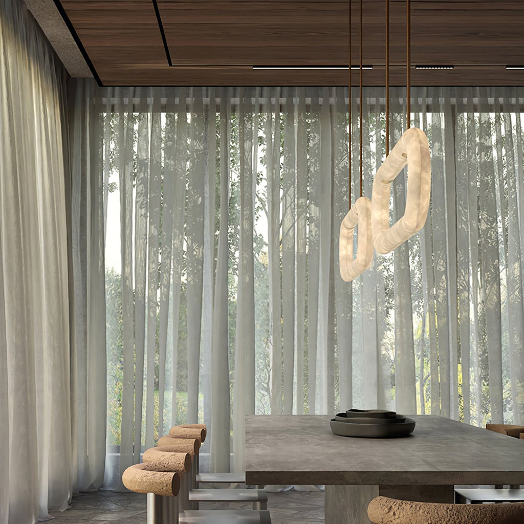 Two Clérachic Vista chandeliers over a concrete dining table, sheer curtains and woodland filling the window behind.