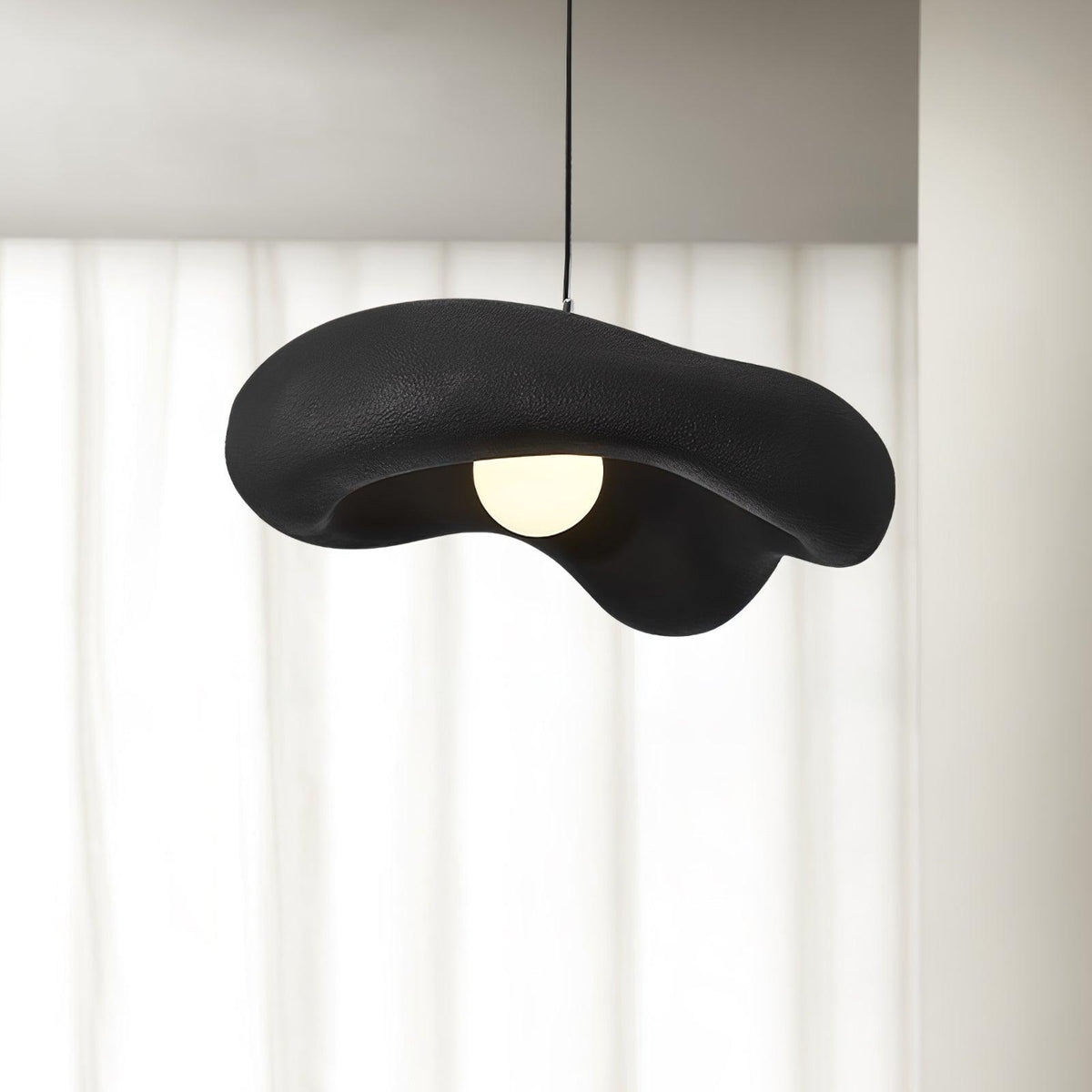 Black Clérachic pendant with a rough, wavy shade and a round bulb, hung in front of sheer white curtains.