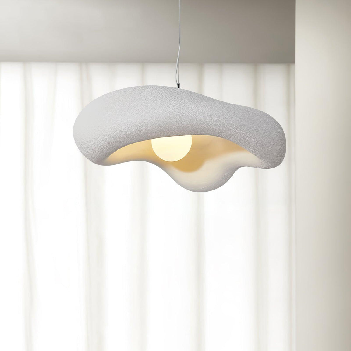 White Clérachic pendant with a rippled, rough-textured shade and a glowing bulb, against sheer white curtains.