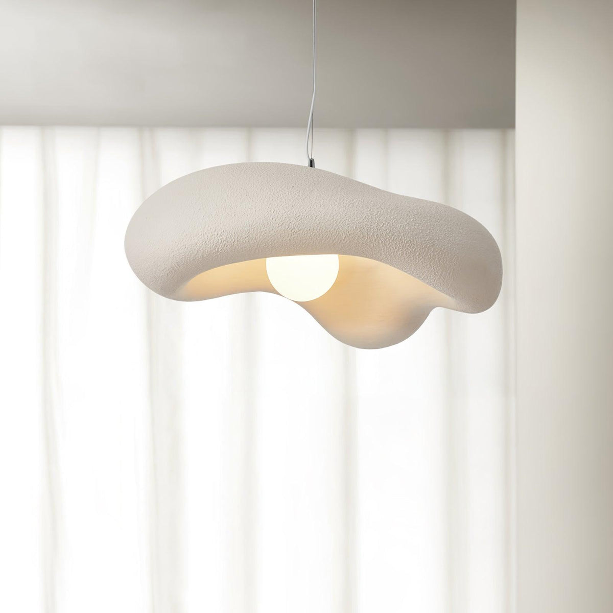Beige Clérachic pendant with a rough, wavy shade and a bare globe bulb, against sheer white curtains.