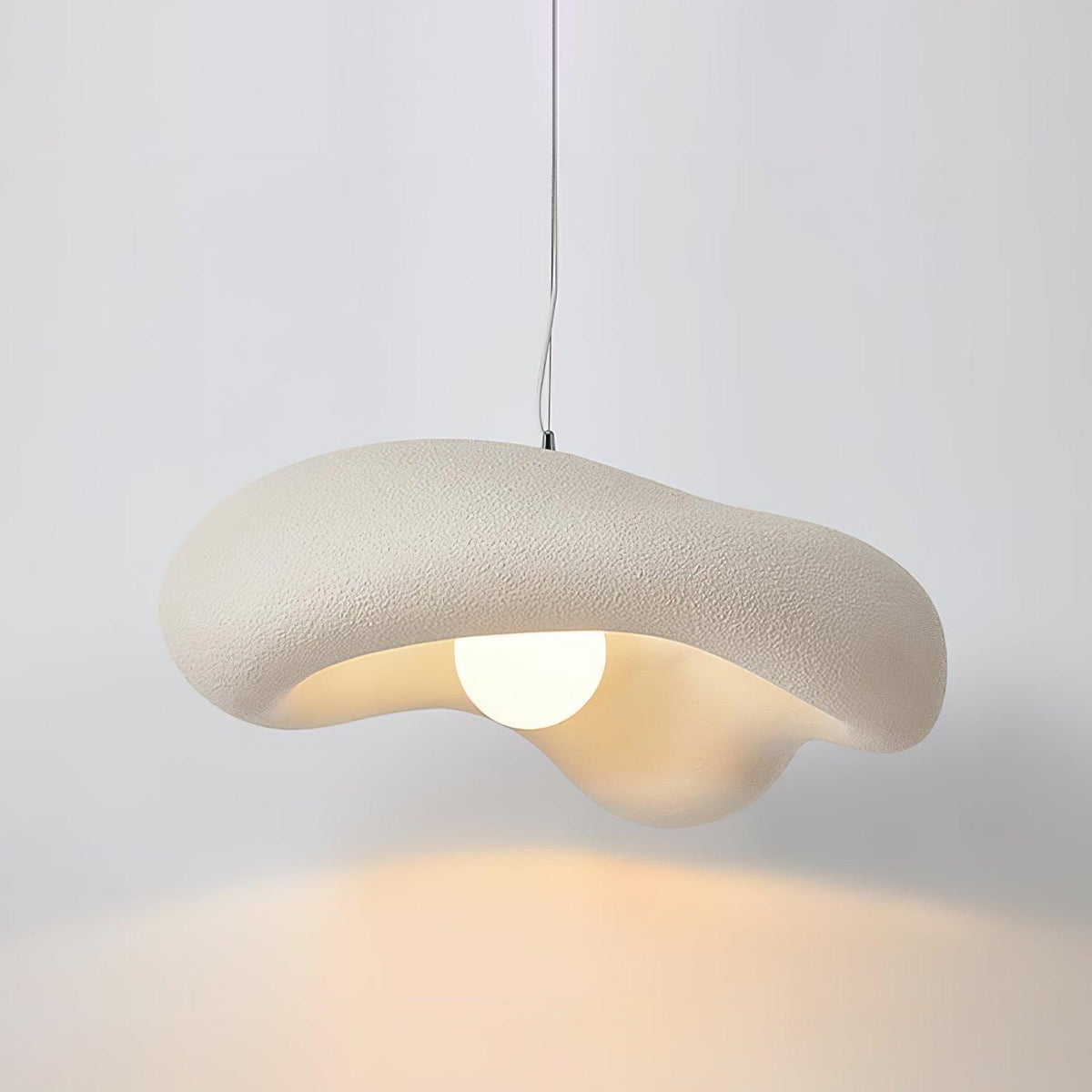 Beige Clérachic pendant lit against a pale grey wall, the shade's wavy edge and a warm pool of light below.