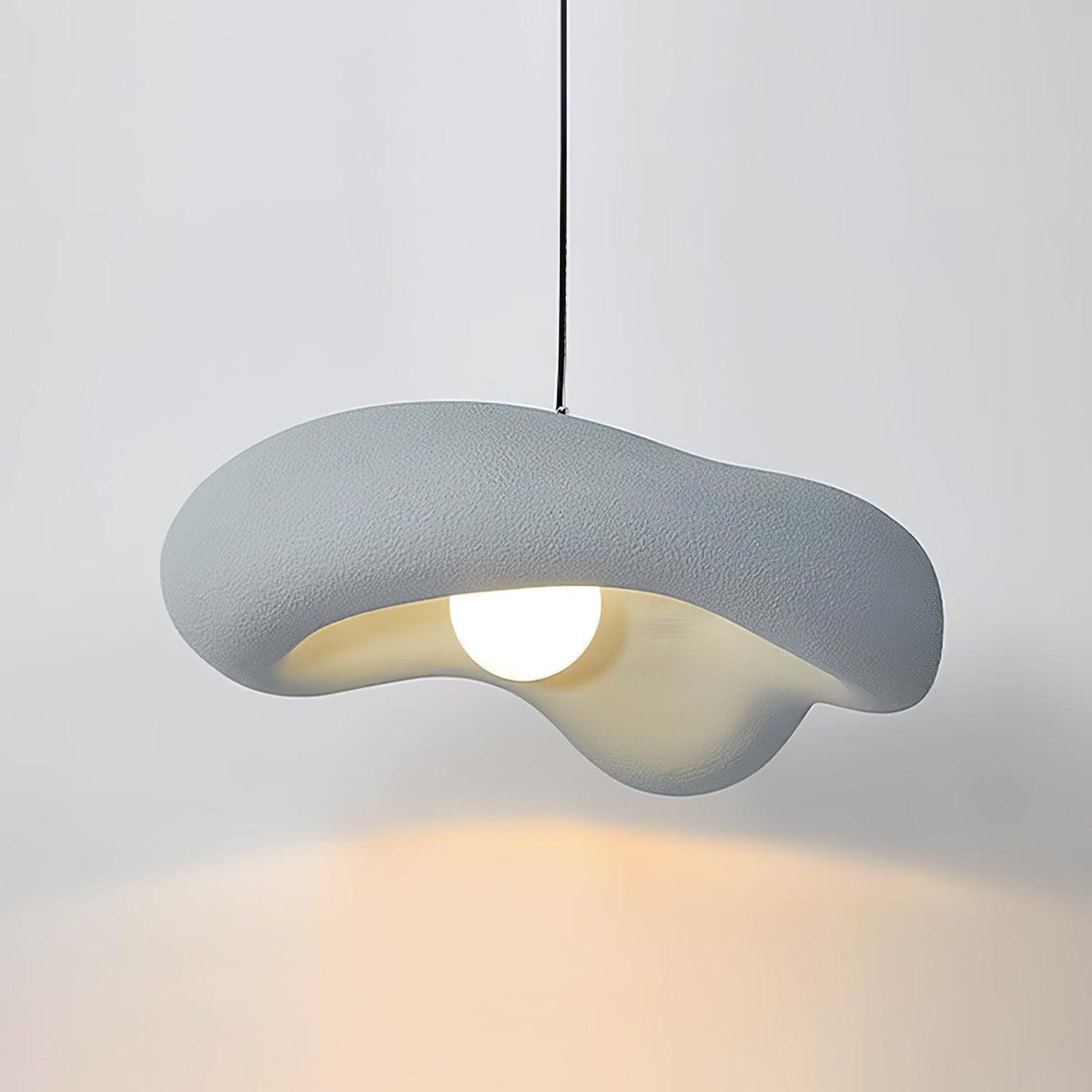 Grey Clérachic pendant lit against a pale wall, the pale blue-grey shade over a round bulb and a warm glow below.